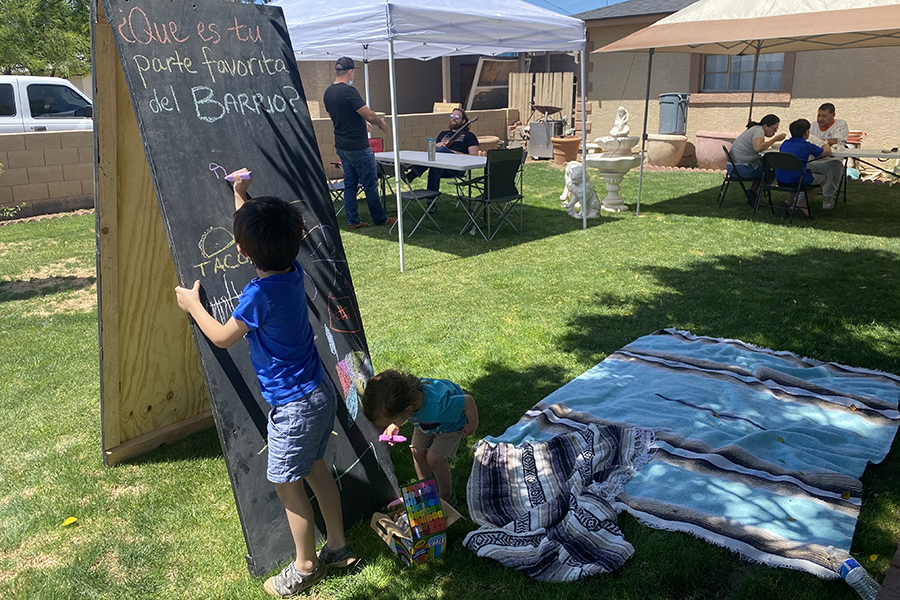 A child writes on a chalkboard that reads "Que es-tu parte favorita del Barrio?" during an outdoor community gathering.